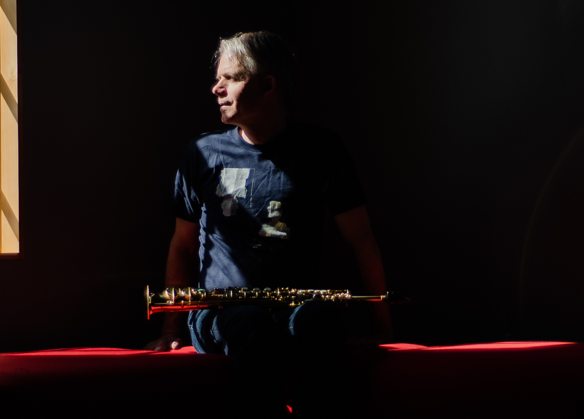 Photo of Titus Abbott with his soprano saxophone on his lap, in a dark space looking towards a sunny window